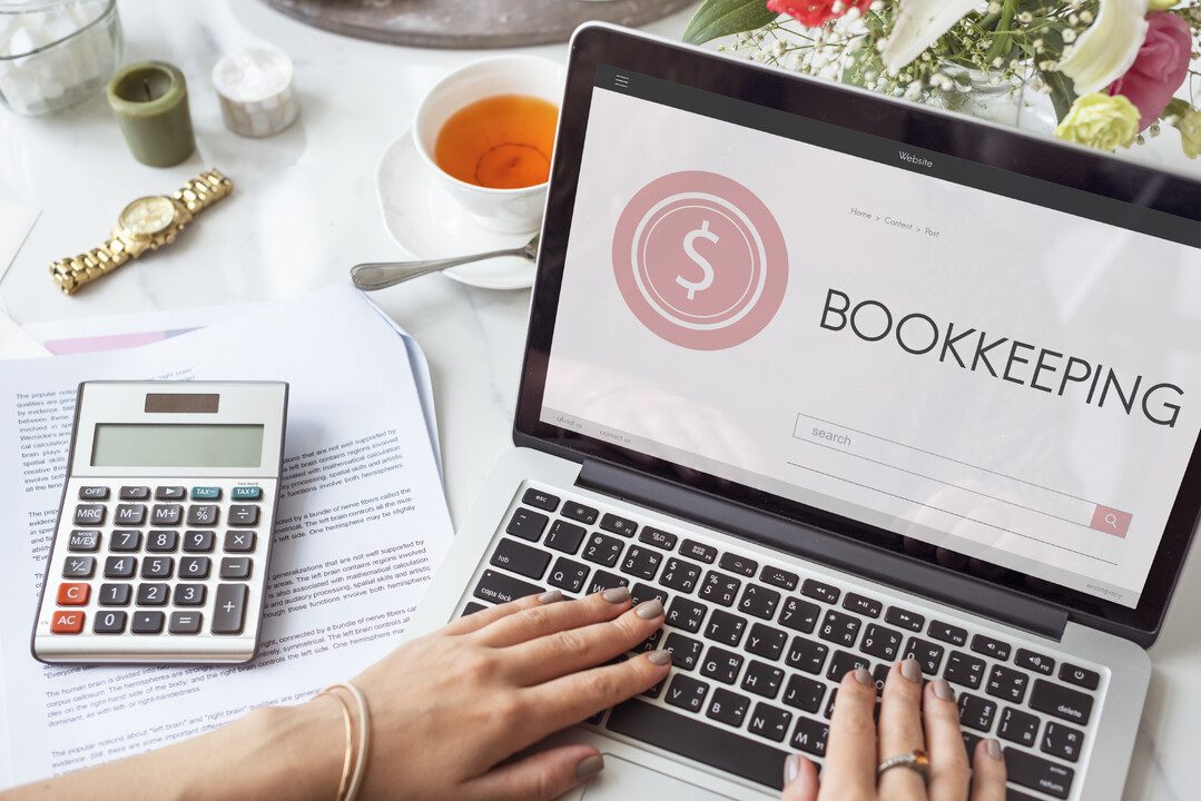 Hands typing on a laptop that shows the word Bookkeeping, next to a calculator and papers